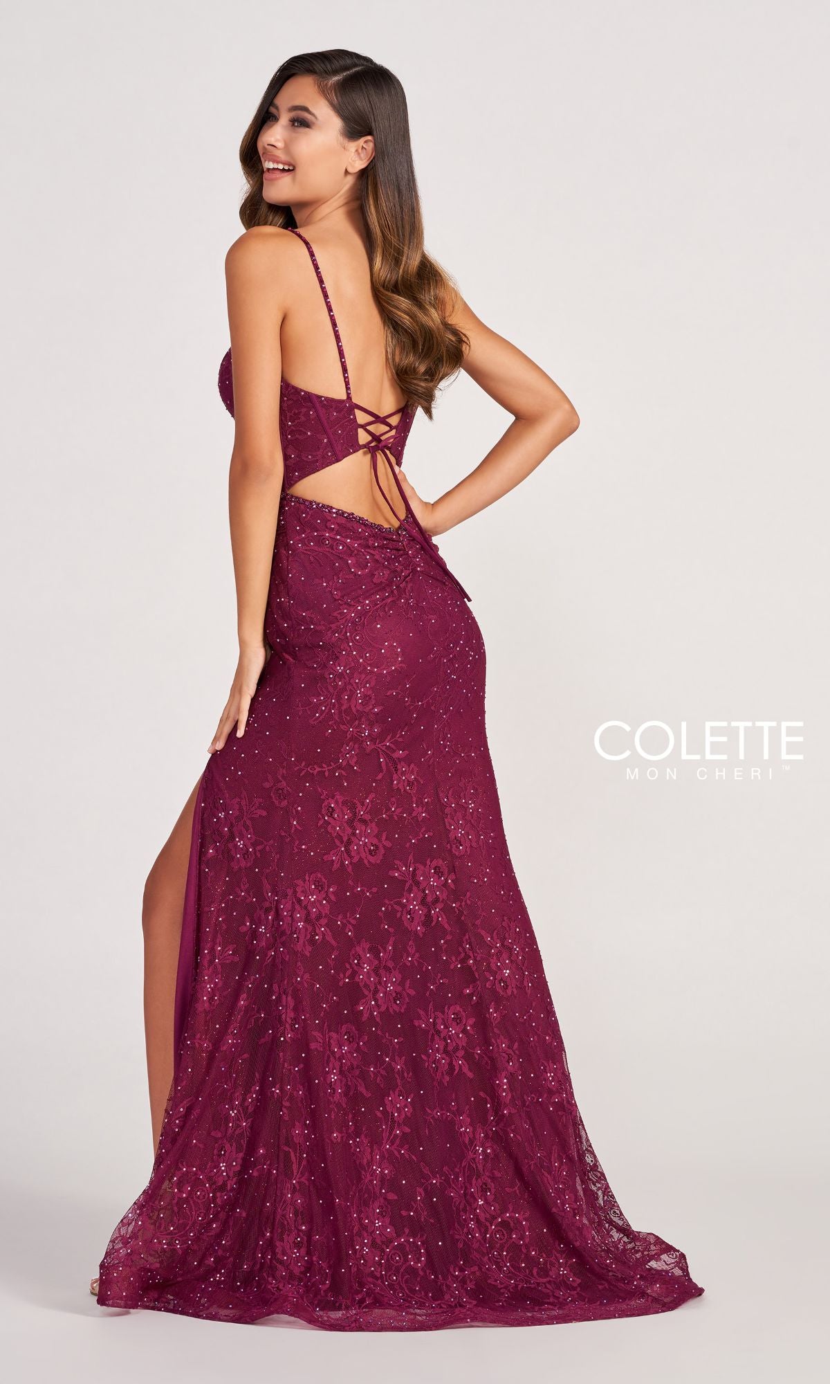 Woman wearing the Colette CL2037 Long Lace Prom Dress with Beads, a floor-length, sleeveless burgundy gown featuring an open back and lace-up detailing, posing and smiling over her shoulder.